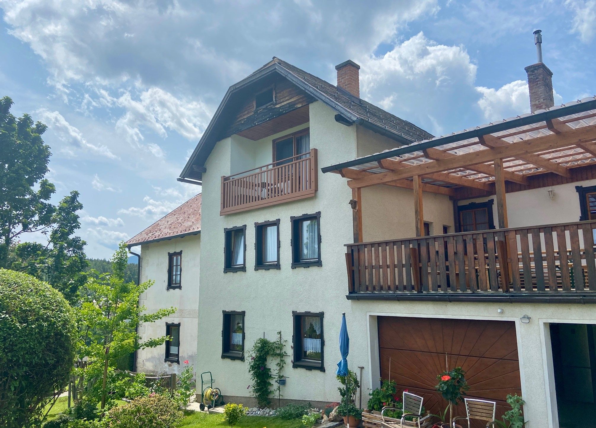 Egy kétszintes ház erkéllyel és terasszal, kerttel és fákkal körülvéve, felhős égbolt alatt.