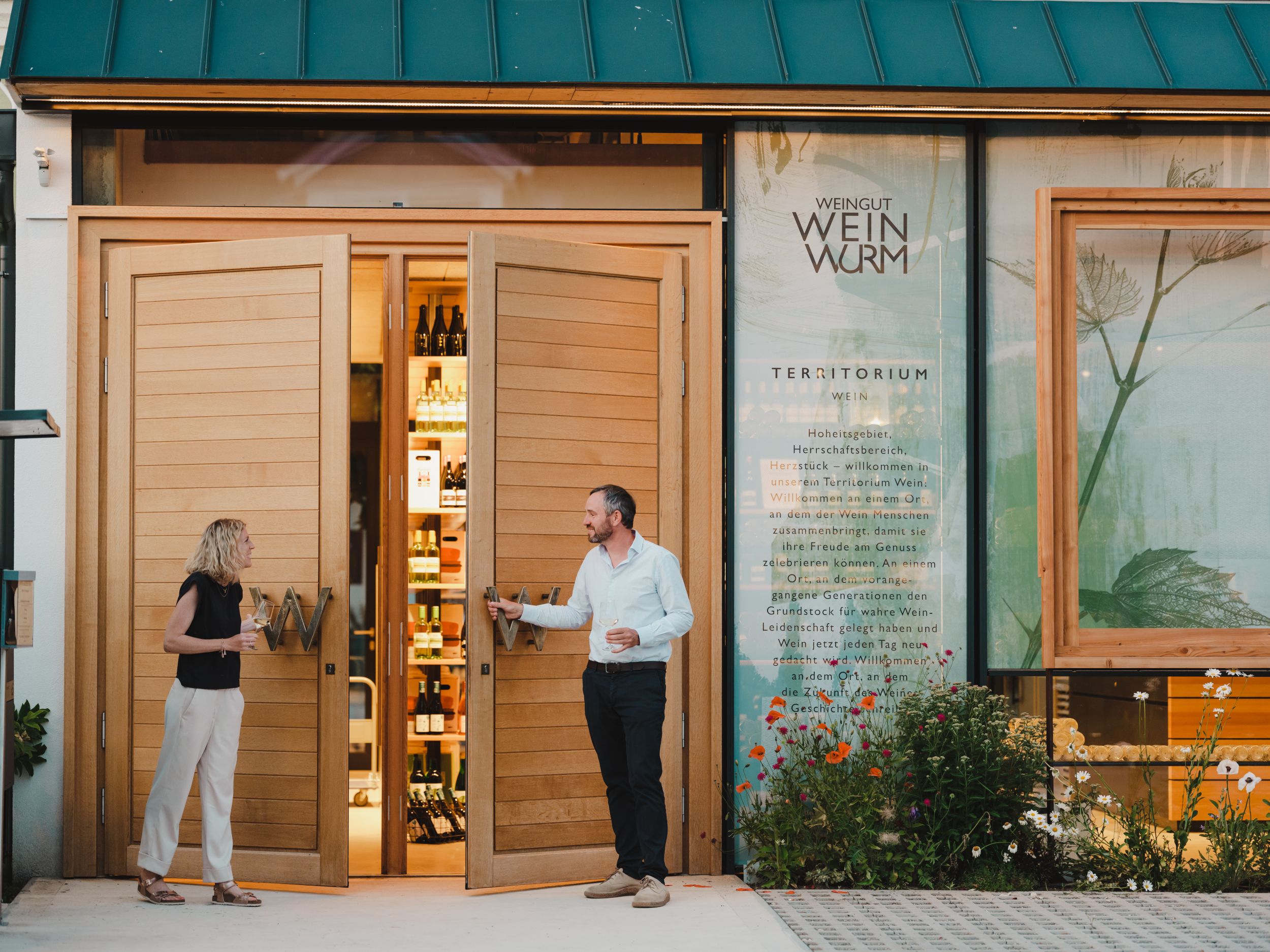 Két ember áll egy borászat előtt, amelynek nyitott faajtói palackokat mutatnak a belsejében.