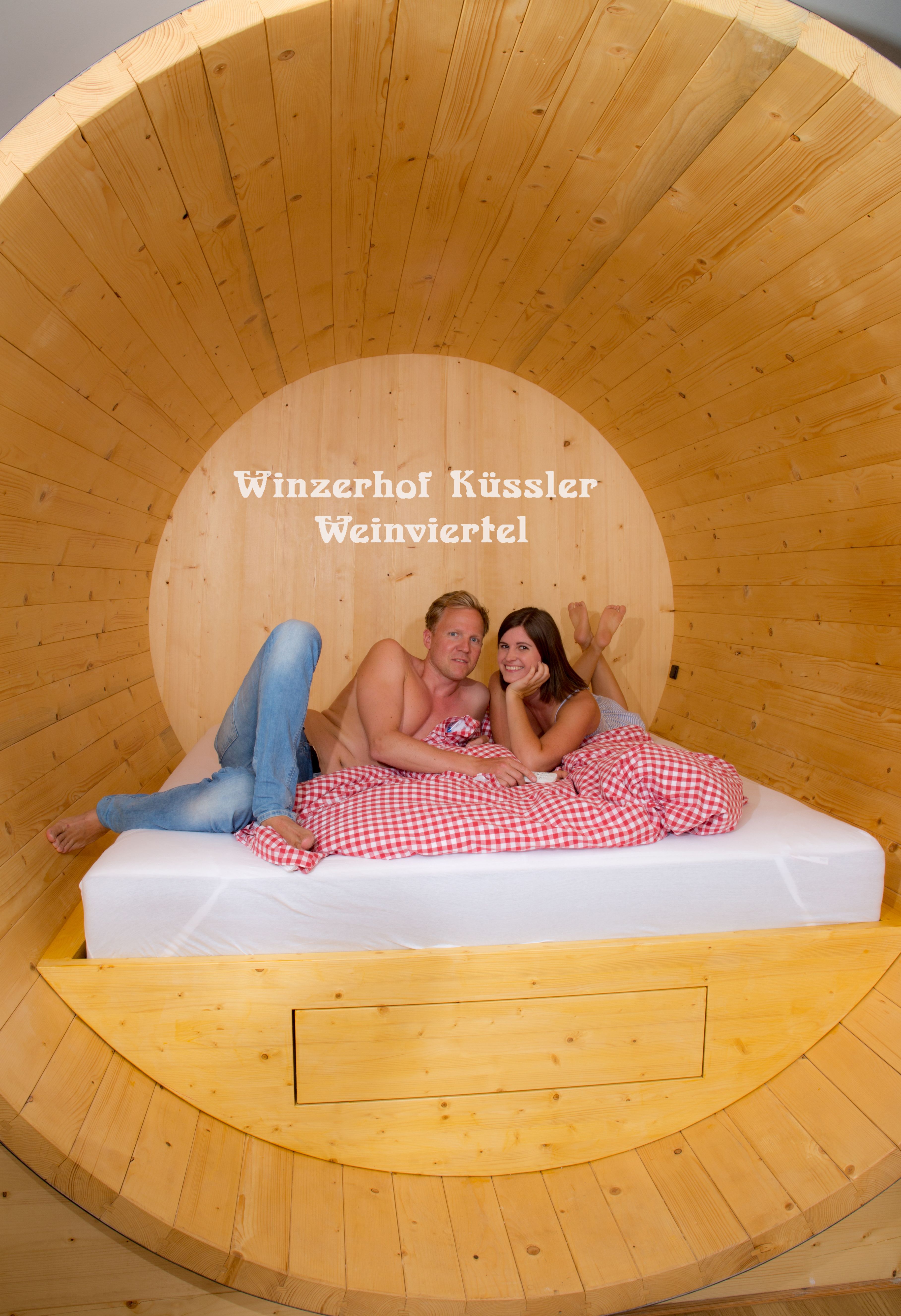 Egy házaspár fekszik egy nagy, piros-fehér kockás ágyneművel borított fa hordóágyban. A háttérben a "Winzerhof Küssler Weinviertel" felirat olvasható.