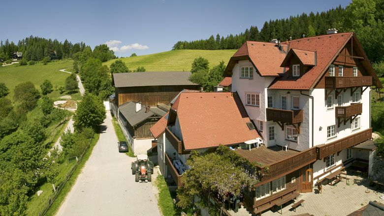 Vörös tetős farm vidéki környezetben, zöld rétekkel és erdőkkel körülvéve.