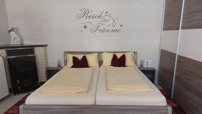 Egy h&aacute;l&oacute;szoba francia&aacute;ggyal, s&aacute;rga &aacute;gyneművel &eacute;s &iacute;r&aacute;ssal a falon. Egy k&aacute;lyha &eacute;s egy kutyaszobor is l&aacute;that&oacute;.