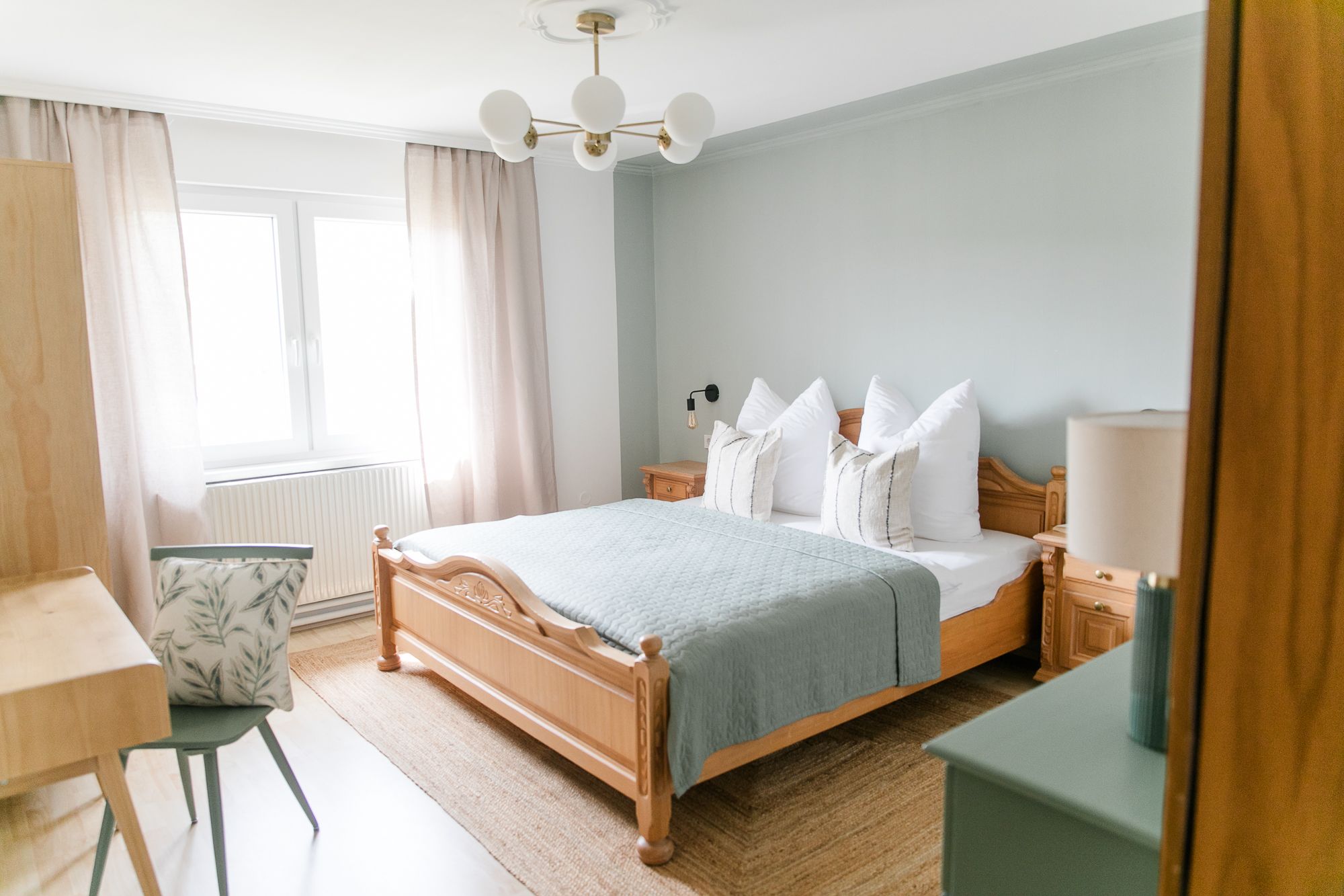 Hangulatos hálószoba faággyal, kék paplannal, éjjeliszekrénnyel és íróasztallal, székkel. Világos színű függönyök és egy ablak látható.