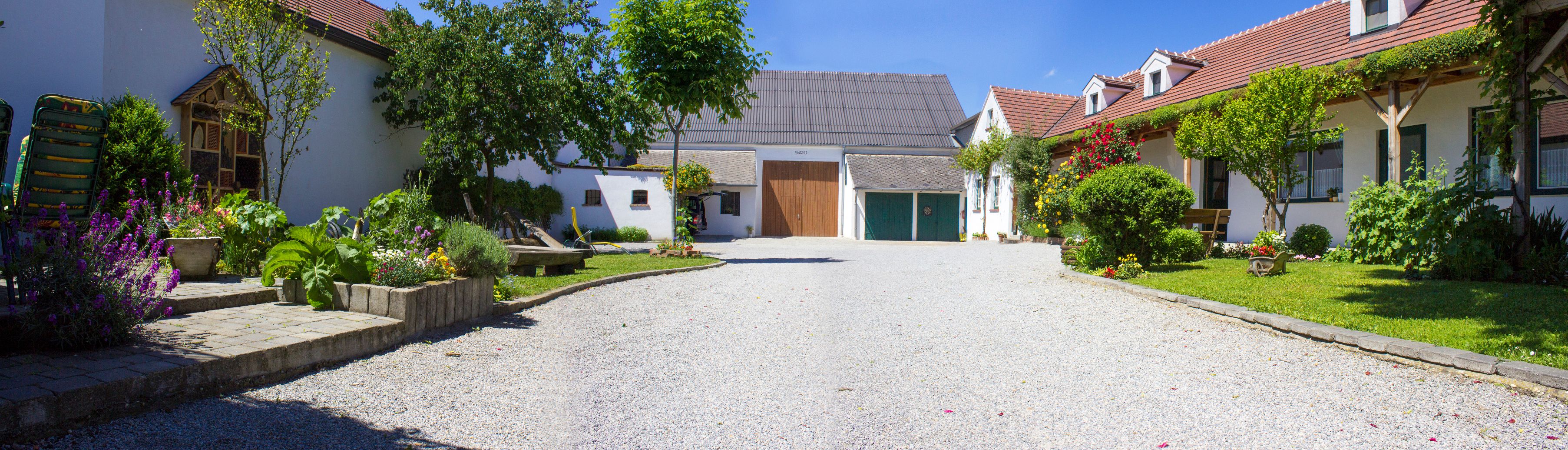 Idilli szőlőskert kavicsos ösvényekkel, virágos kertekkel és hagyományos épületekkel a kék ég alatt.