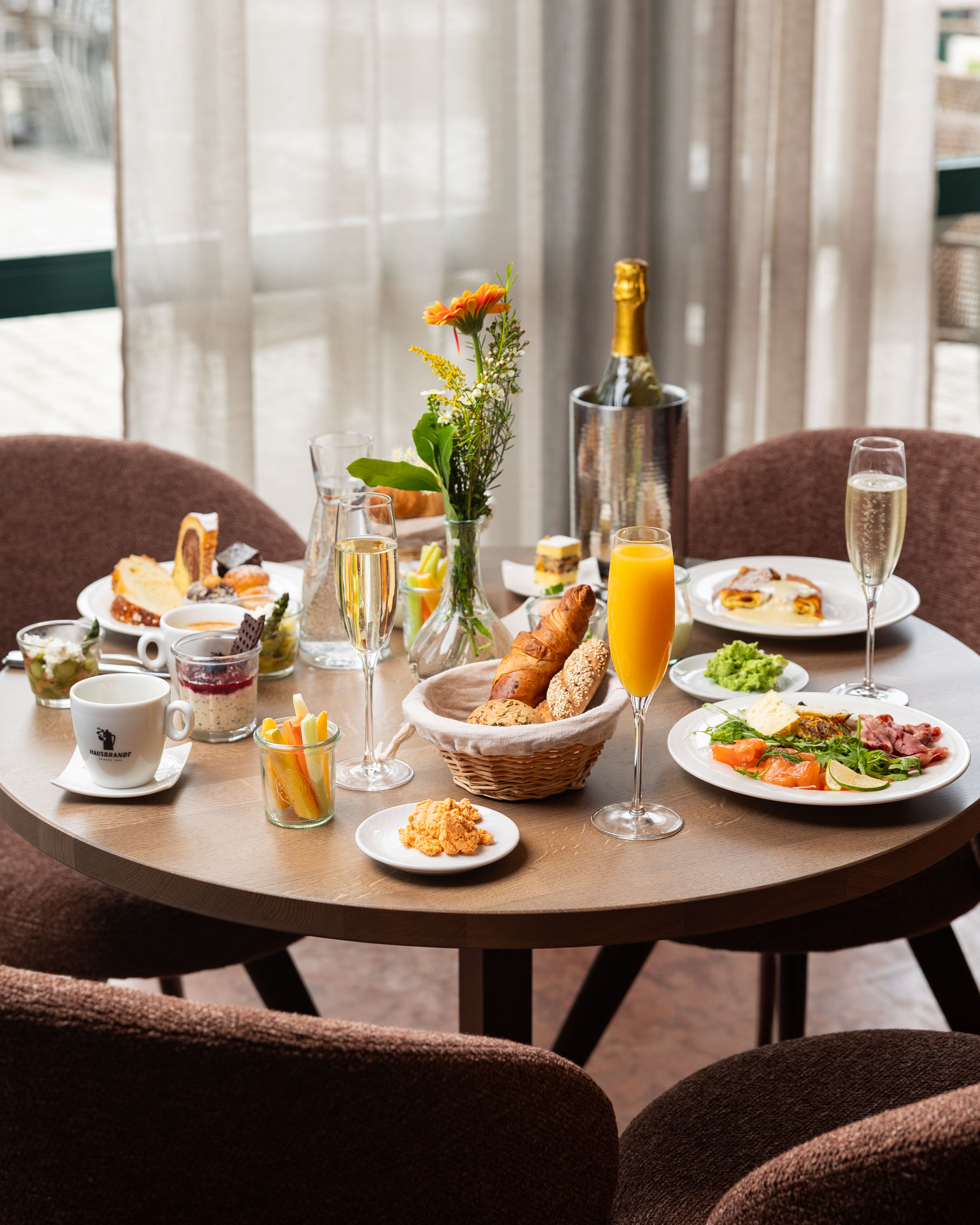 Megterített reggelizőasztal különböző ételekkel, italokkal és virágokkal.