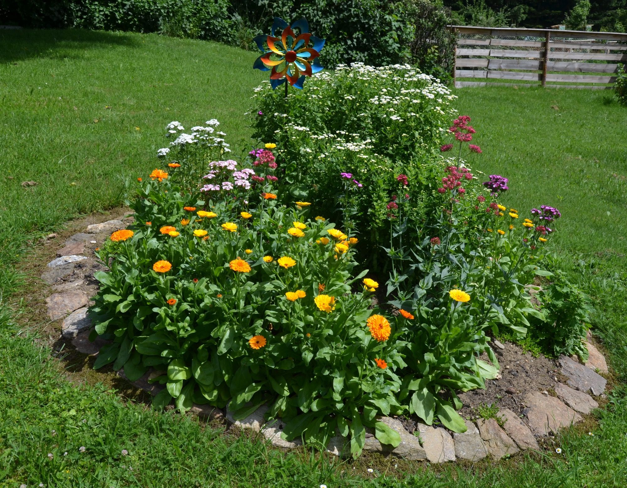 Kerek virágágyás színes virágokkal és egy szélmalom a kertben.