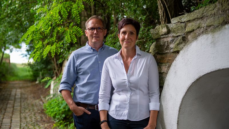 Egy férfi és egy nő áll egymás mellett egy kövezett ösvényen a szabadban, fákkal és kőfallal körülvéve.