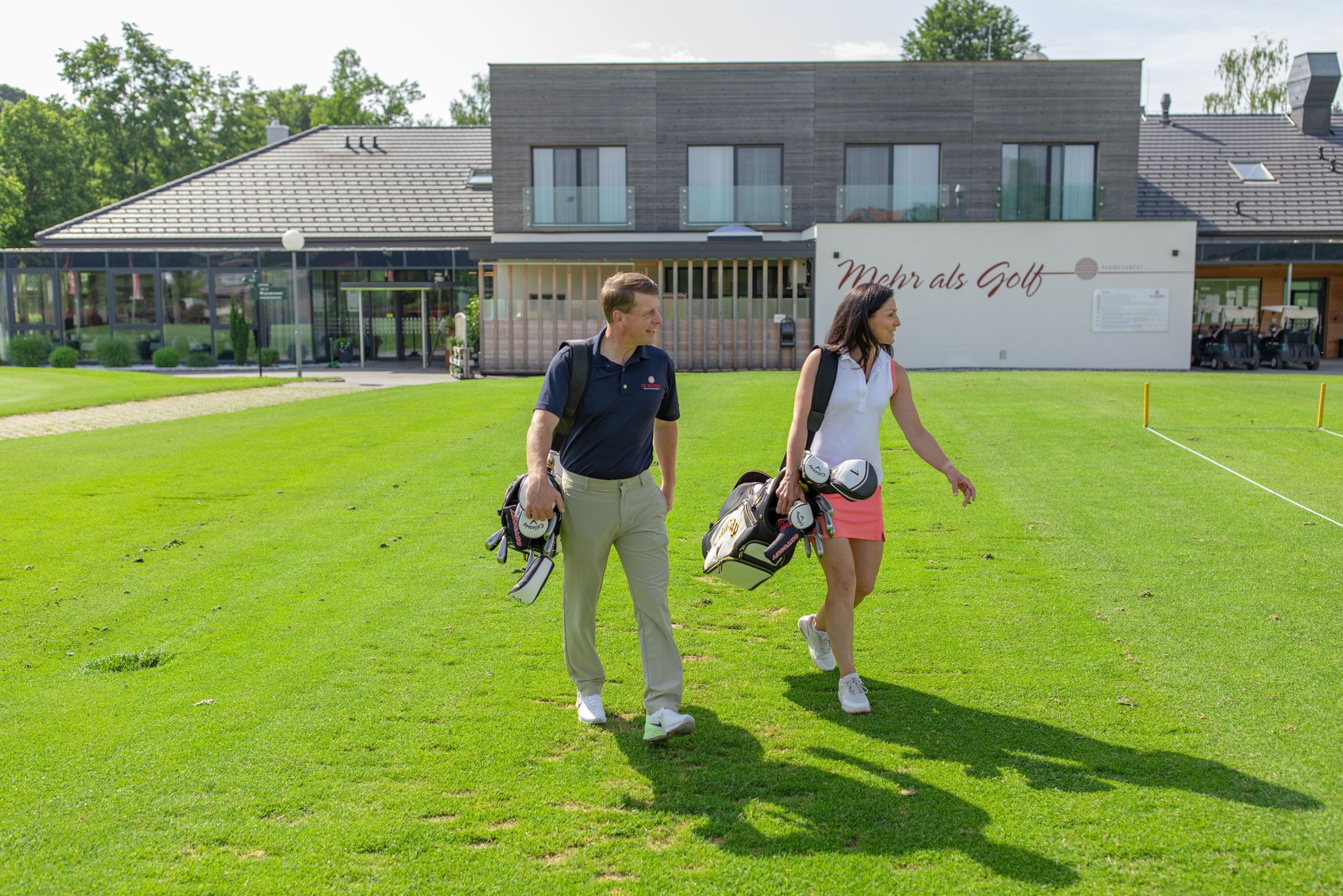 Két ember golftáskával egy modern klubház előtt.