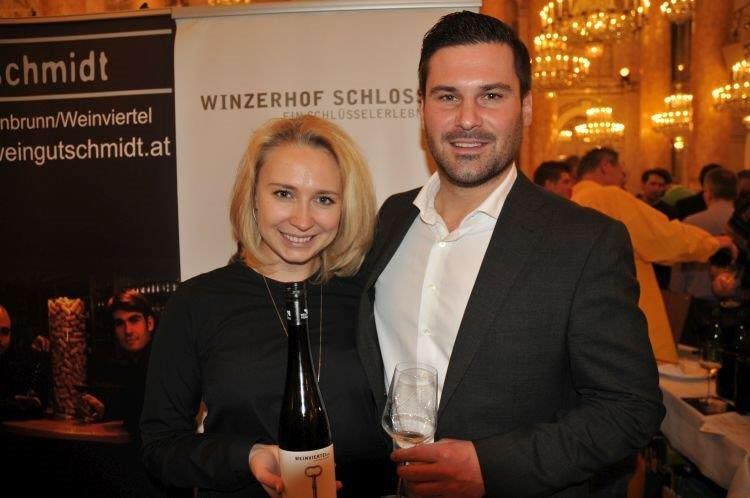 Egy házaspár egy borosüveget és egy borospoharat tart a "Winzerhof Schlosser" feliratú transzparens előtt.