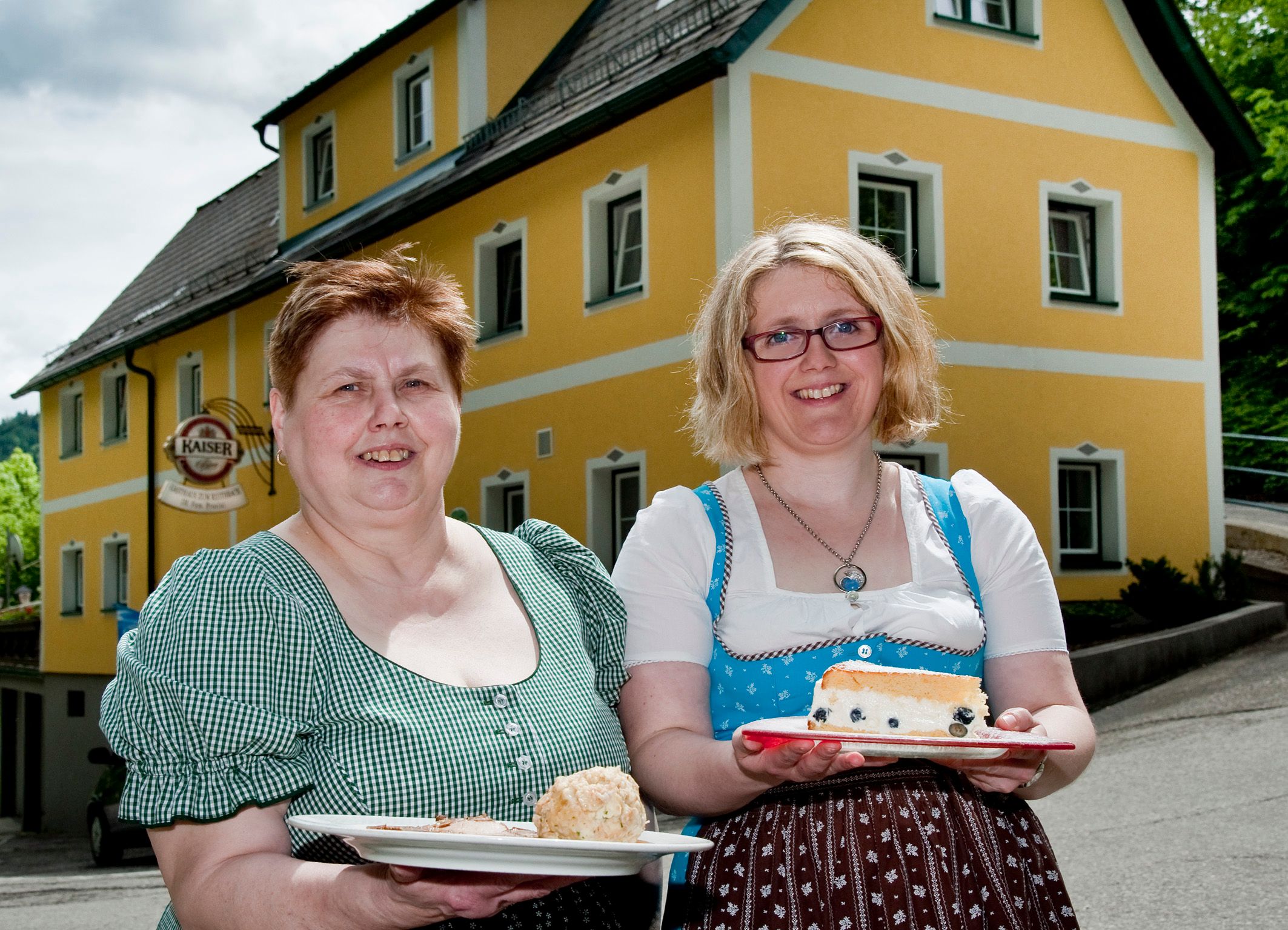 Két hagyományos ruhás nő tart tányérnyi ételt egy sárga épület előtt.
