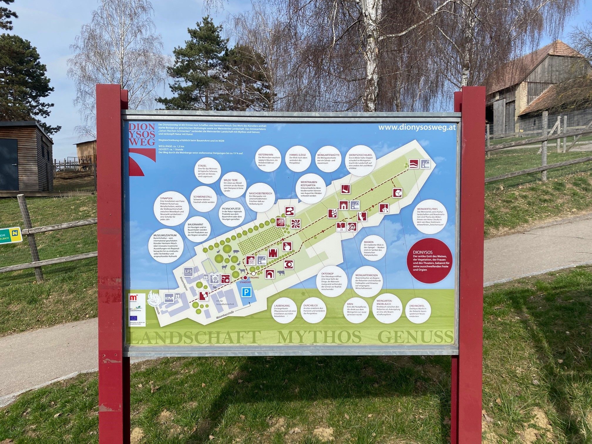 A Dionüszosz-ösvény információs táblája térképével és magyarázatokkal, fákkal és épületekkel körülvéve.