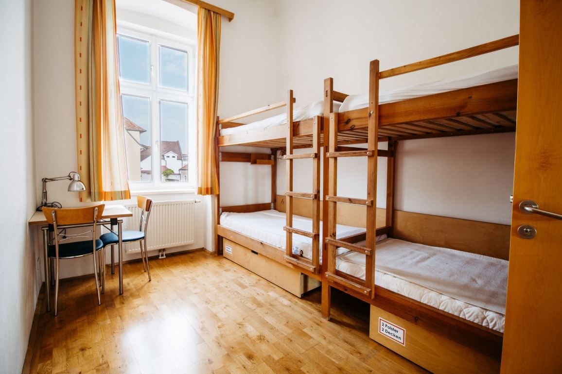 Egyszerű szoba emeletes ággyal, asztallal és székkel, világos függönyökkel az ablakon.