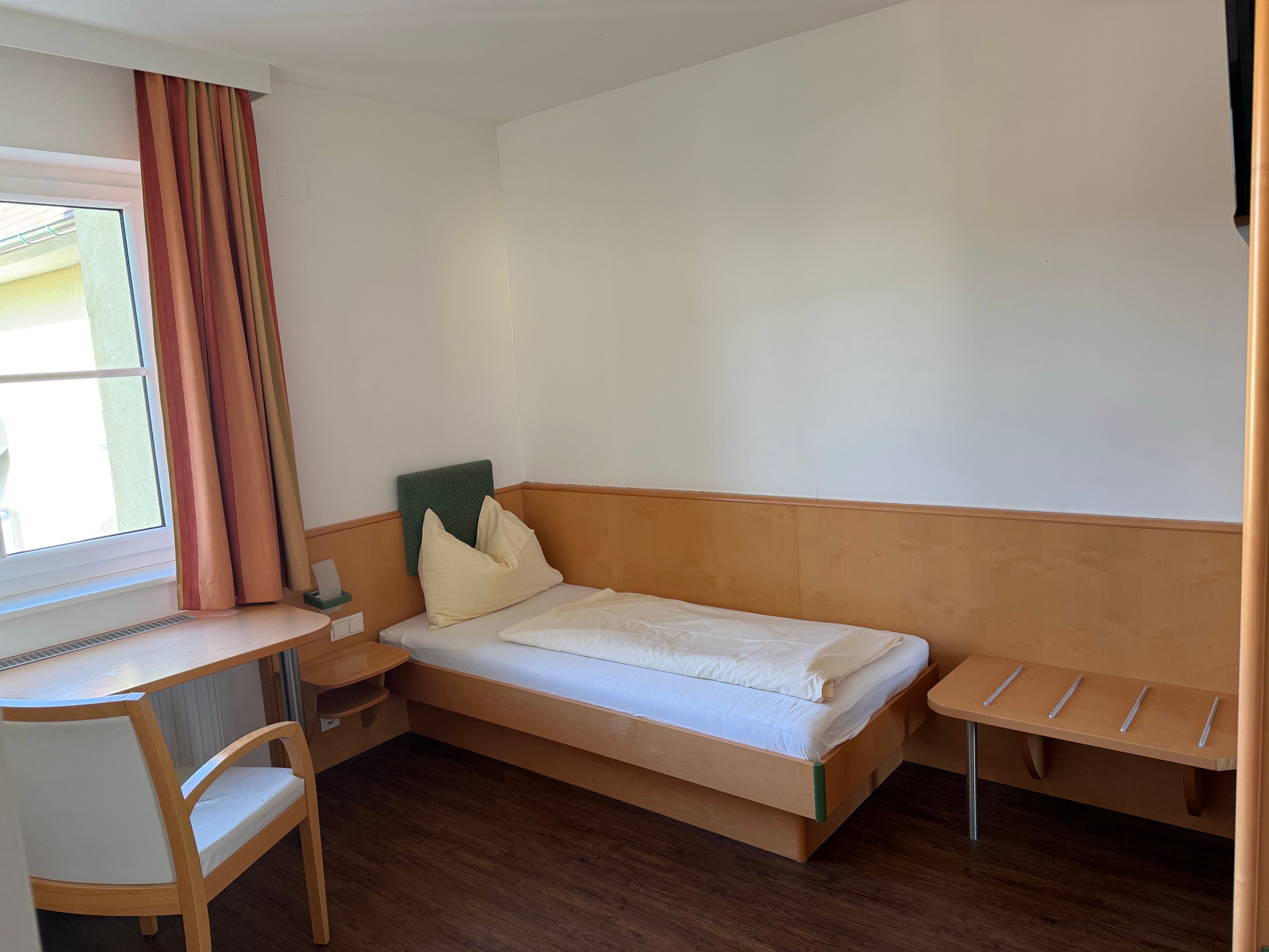 Egy egyszerű szállodai szoba egy egyszemélyes ággyal, asztallal, székkel és függönyös ablakkal.