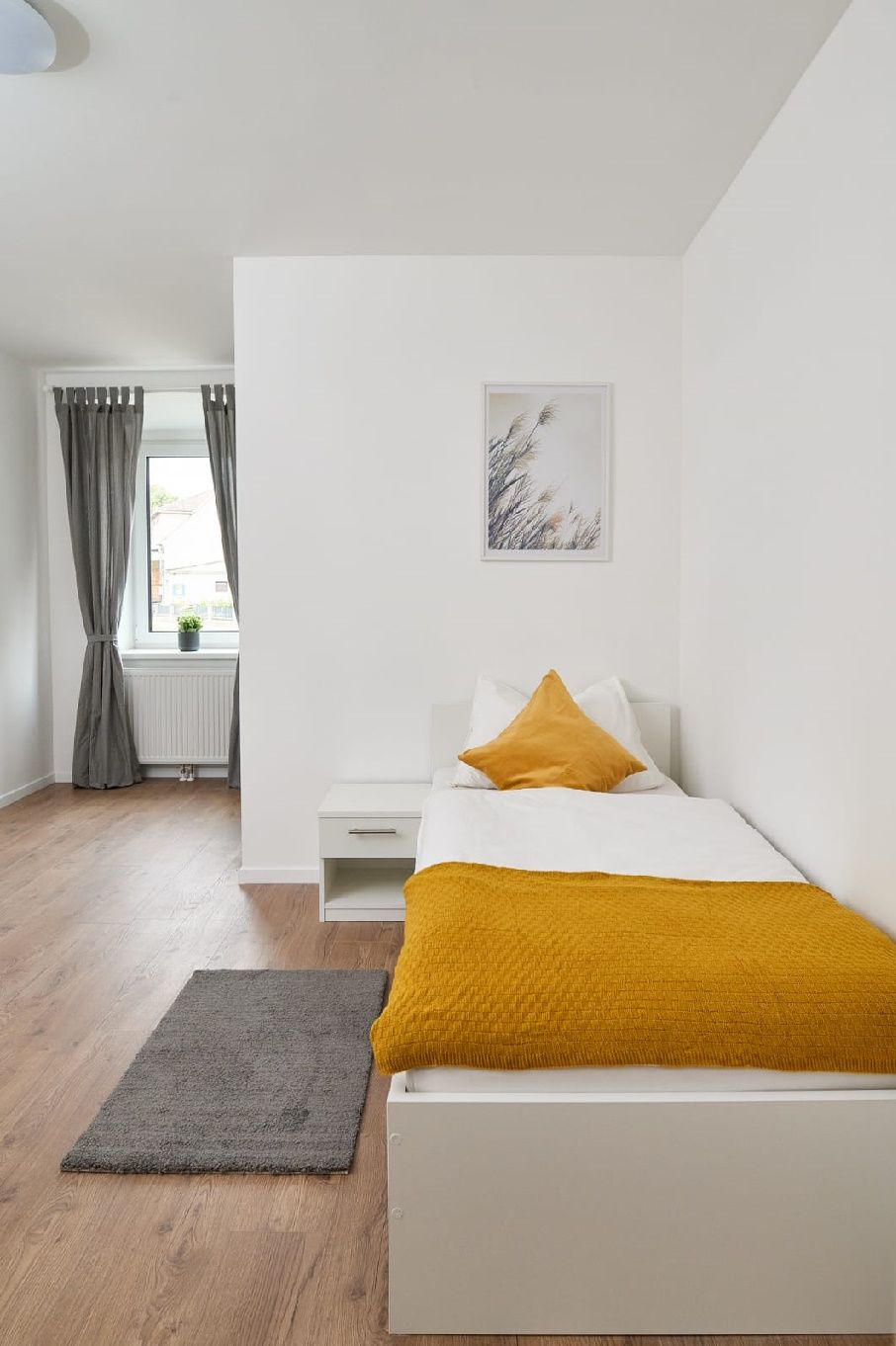 Egyágyas szoba ággyal, éjjeliszekrénnyel és függönyös ablakkal.