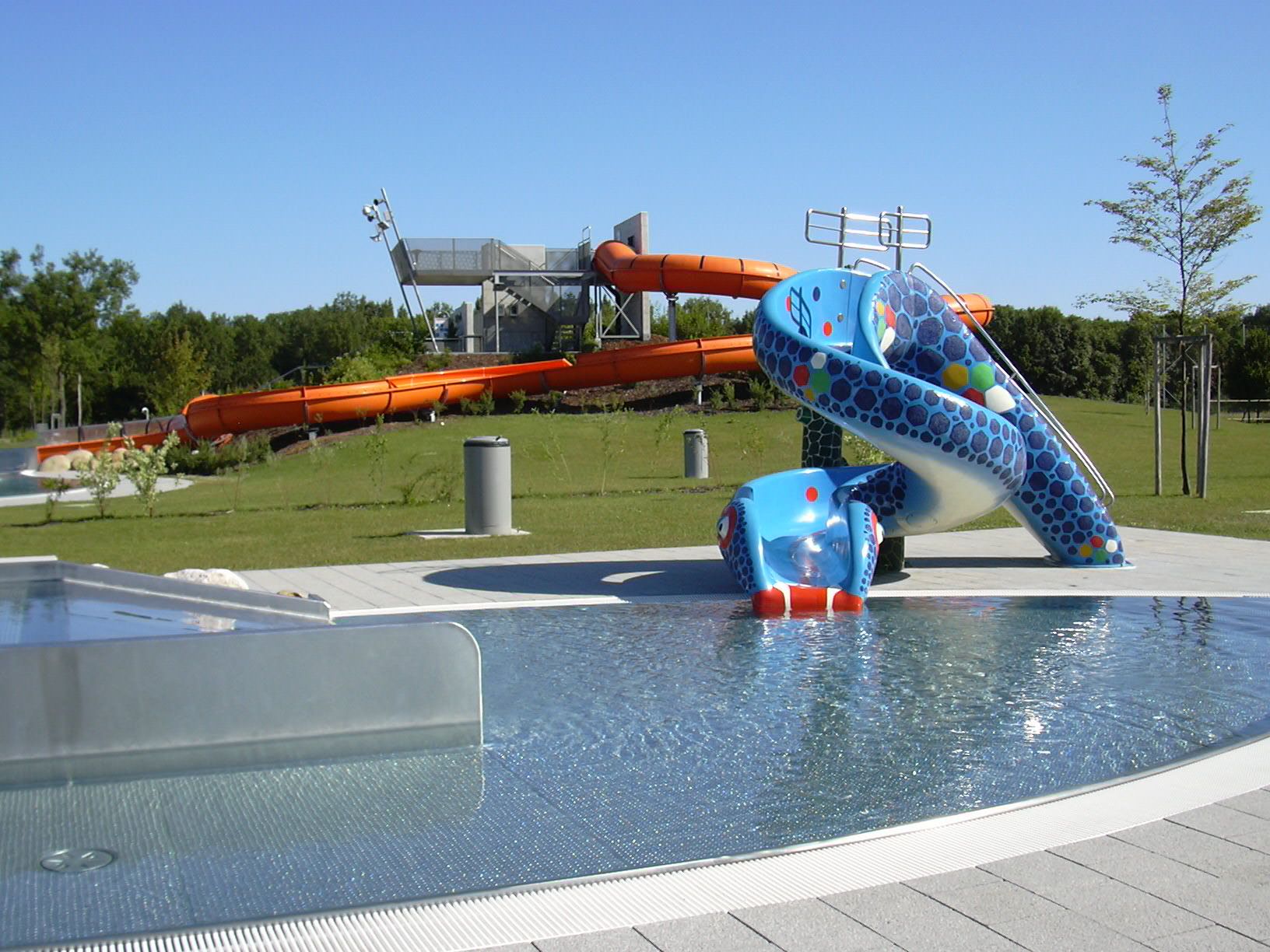 Egy aquapark látképe, a háttérben egy kék gyermekcsúszdával és egy nagy narancssárga vízicsúszdával.