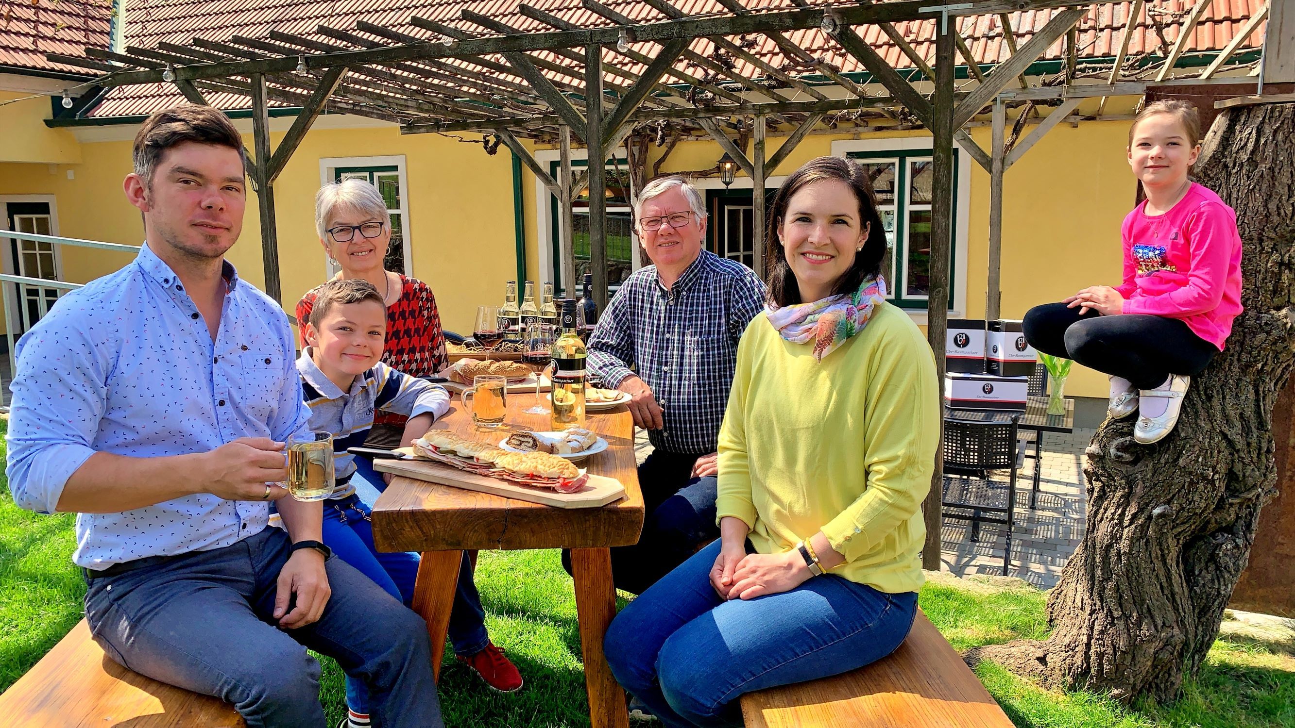 Egy család ül egy fából készült asztalnál a szabadban, és étkezik. Egy gyermek egy fatörzsön ül. A háttérben egy sárga ház pergolával.