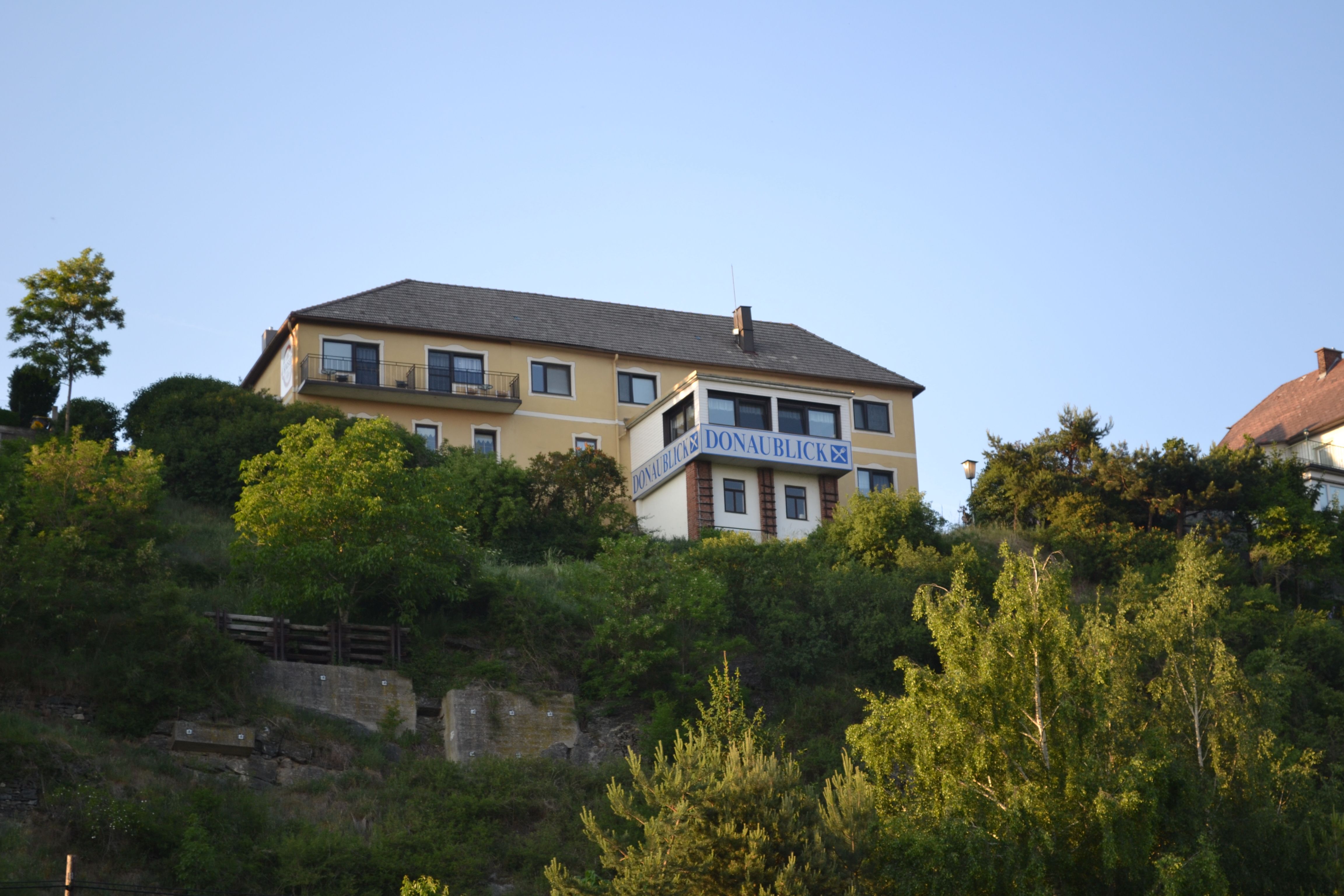 Egy sárga, "Donaublick" feliratú épület egy dombon, fákkal és kék éggel körülvéve.