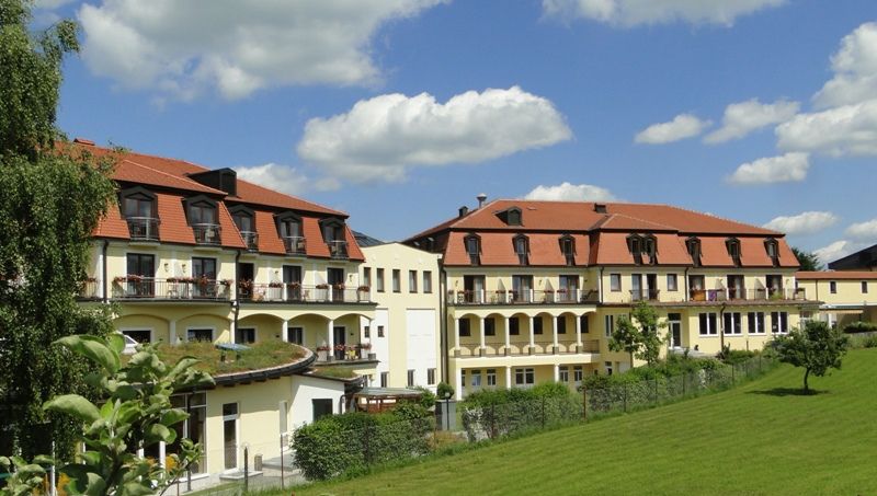 Kurhotel Moorbad Großpertholz piros tetővel és erkélyekkel, zöld rétekkel és fákkal körülvéve, kék ég alatt.