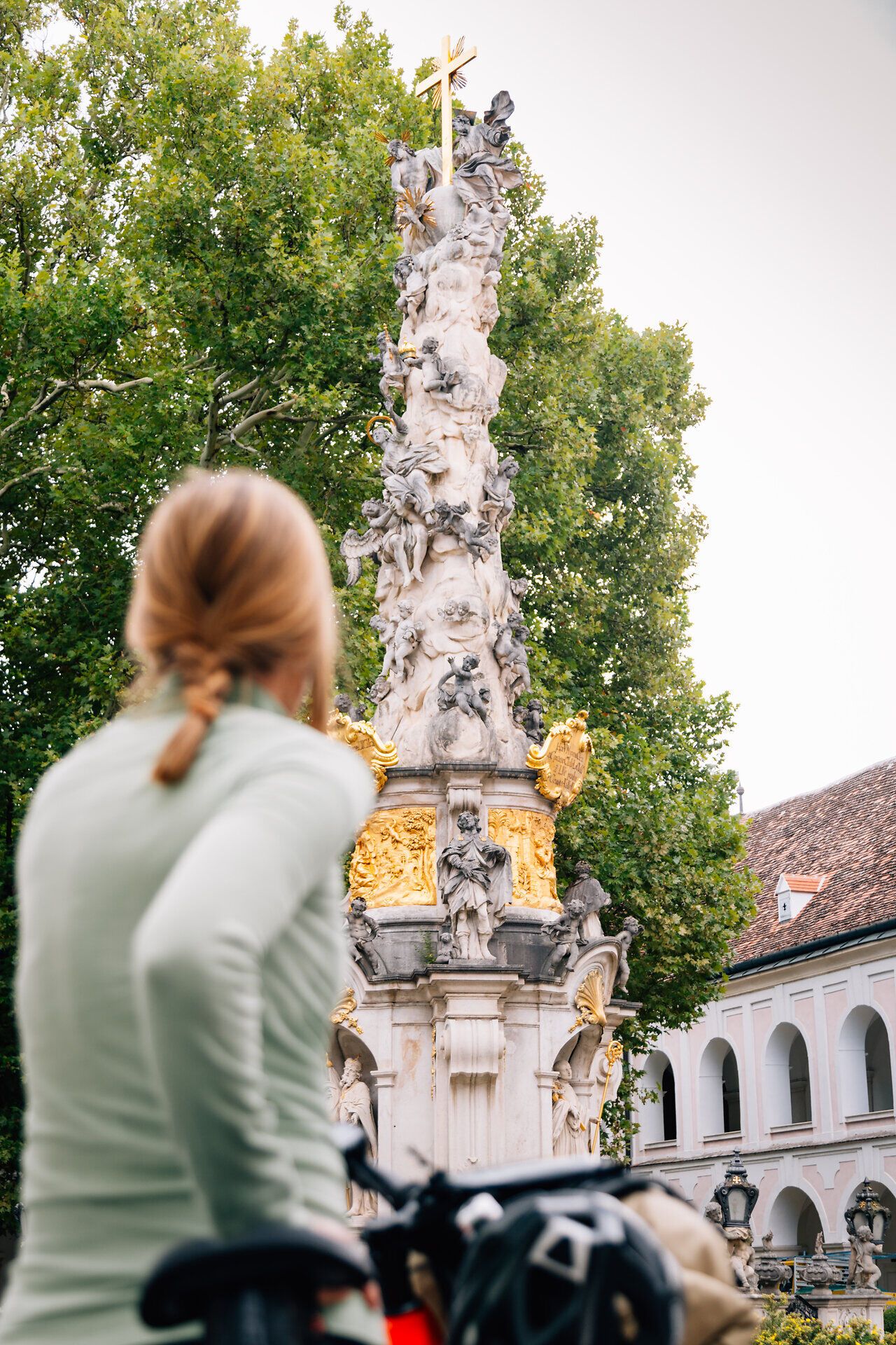 Der Klosterhof Heiligenkreuz strahlt eine friedliche Atmosphäre aus, umgeben von majestätischen Bäumen und kunstvollen Statuen. Radfahrer genießen die frische Luft und die malerische Kulisse, während sie die Schönheit des Wienerwaldes erkunden.