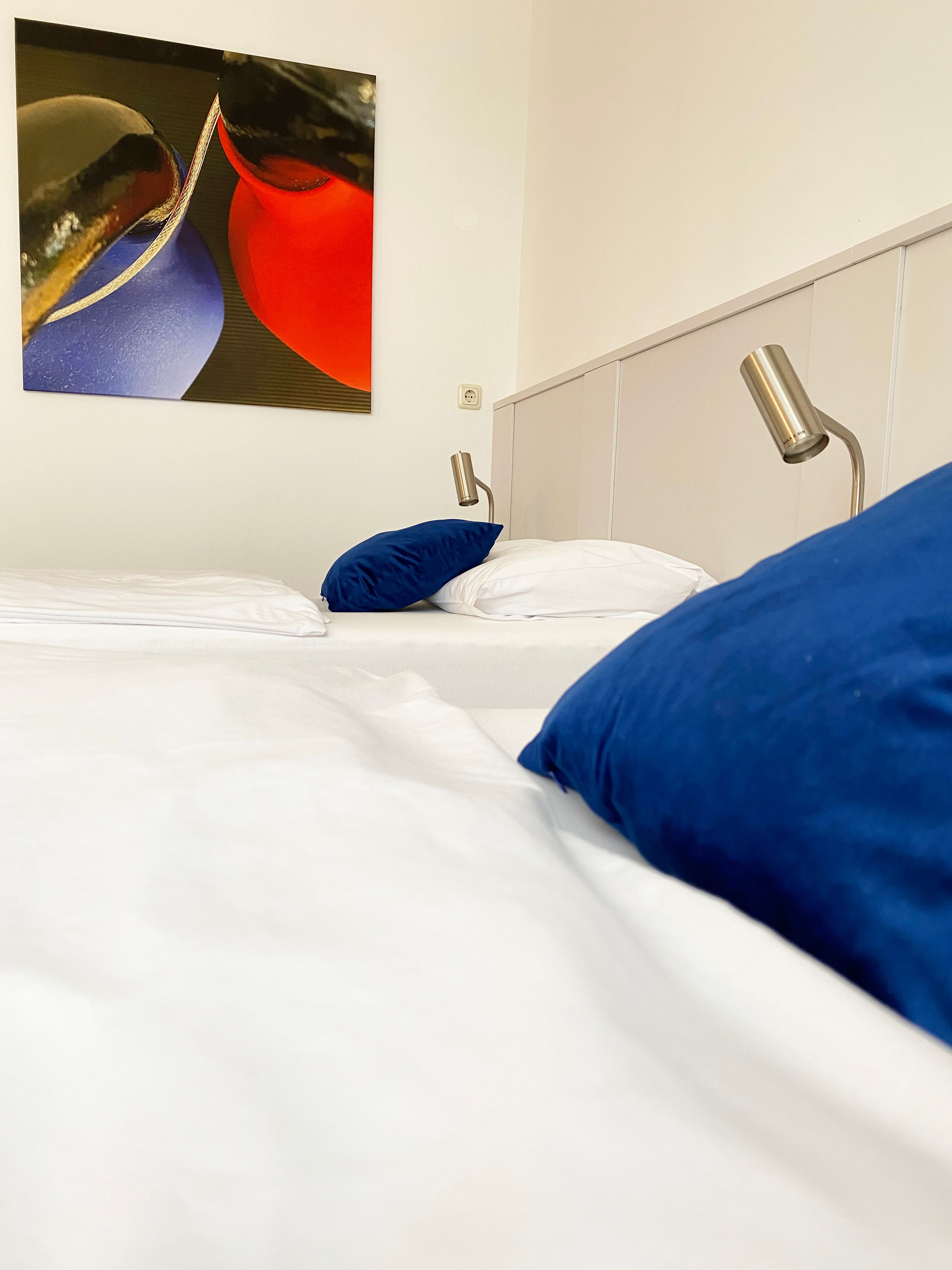 Egy vendégszoba egy ággyal, kék párnákkal, olvasólámpákkal és egy absztrakt festménnyel a falon.