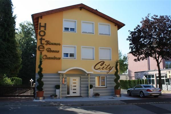 "City Hotel" feliratú sárga épület, körülötte fák és egy parkoló autó.