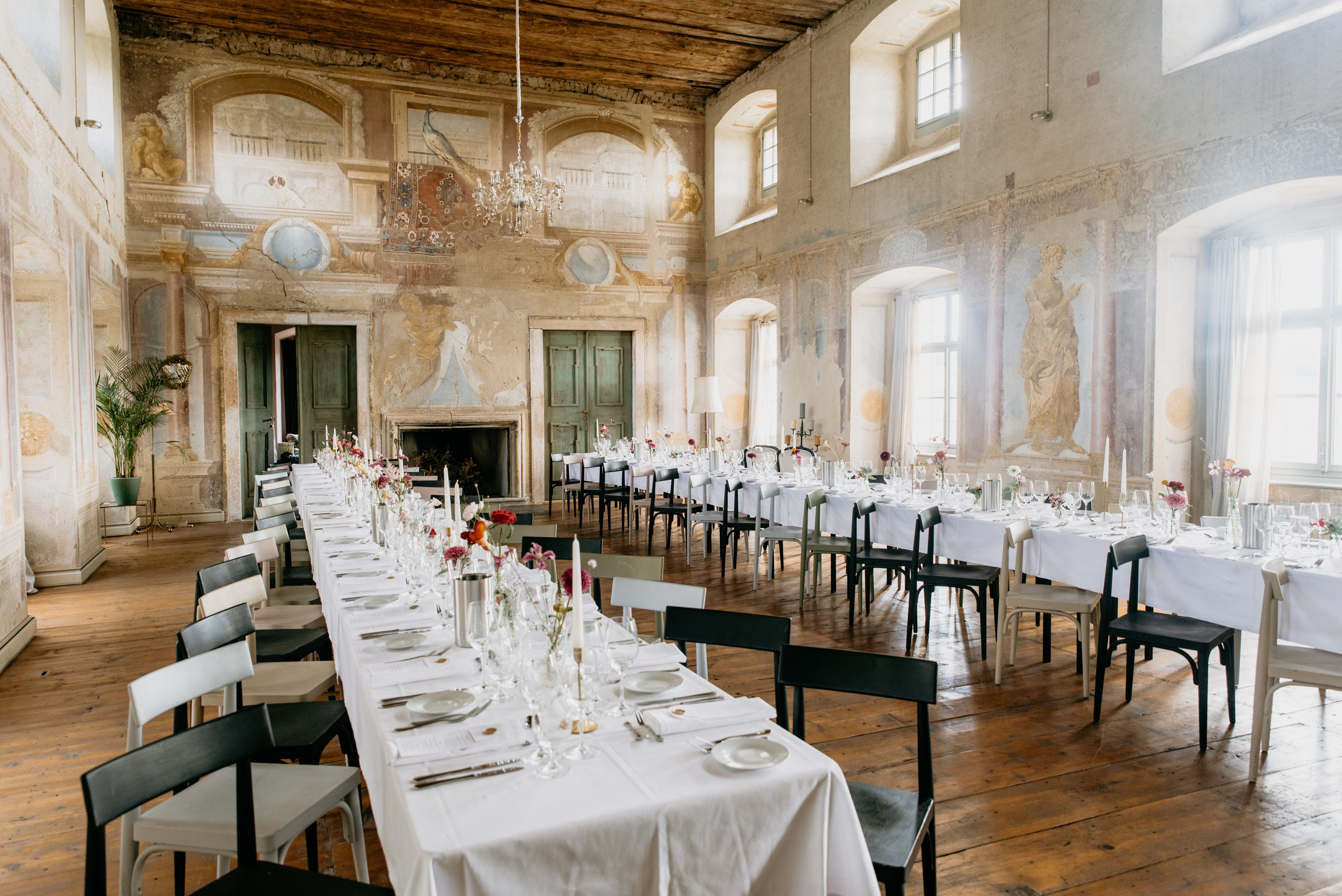 Elegáns díszterem hosszú, terített asztalokkal, antik falfestményekkel és csillárokkal.