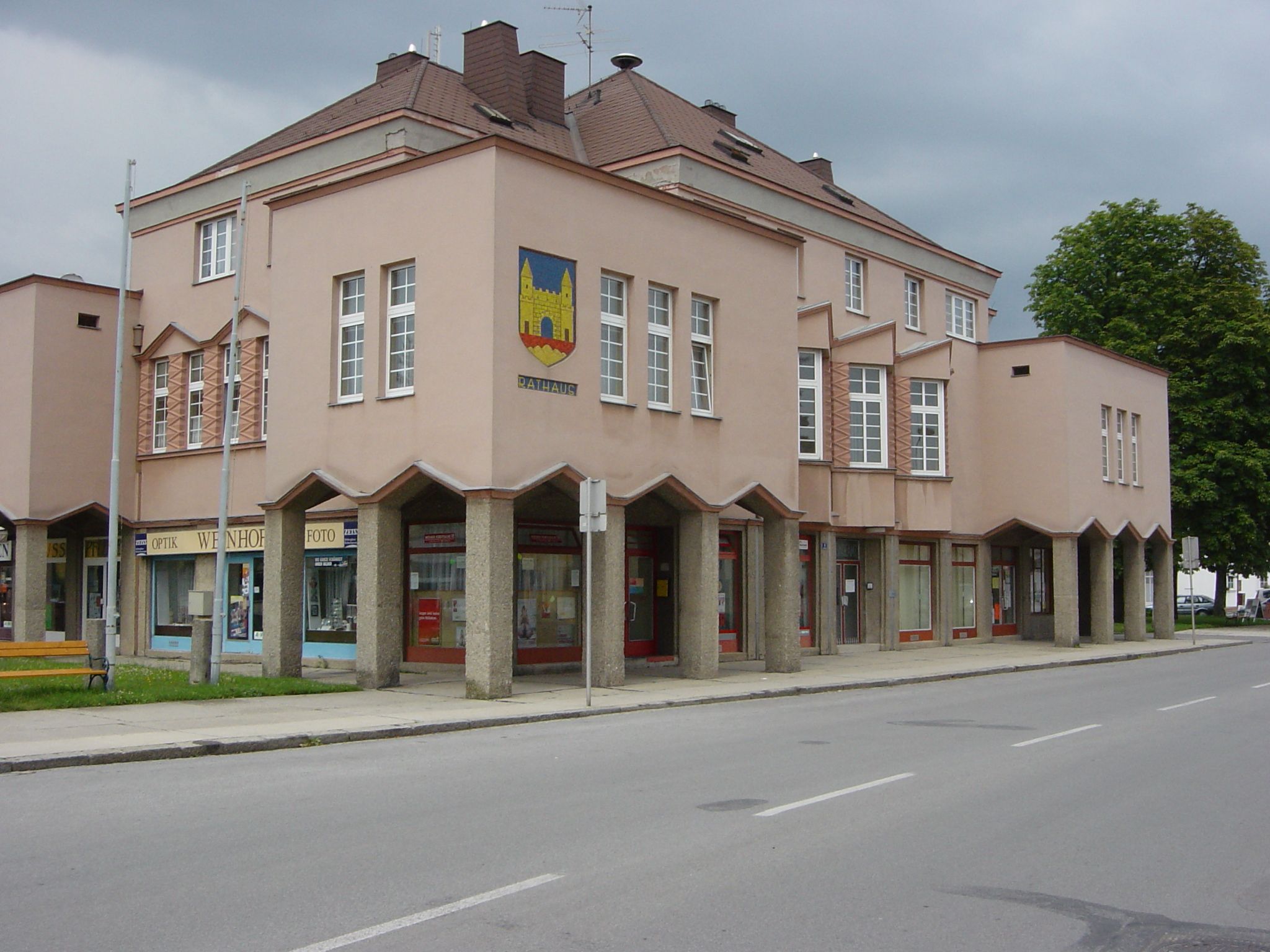 A modern városháza épülete rózsaszín homlokzattal és nagy ablakokkal. A sarkon címer látható, a földszinten pedig üzletek.