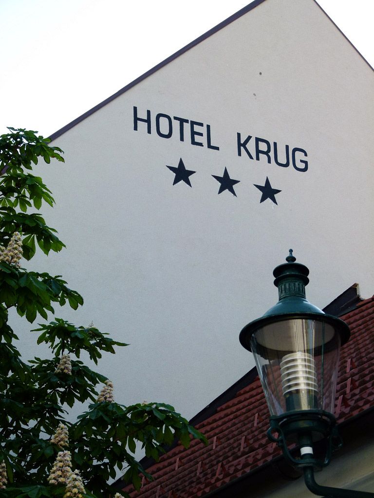 A Hotel Krug homlokzata három csillaggal, fával és utcai lámpával.