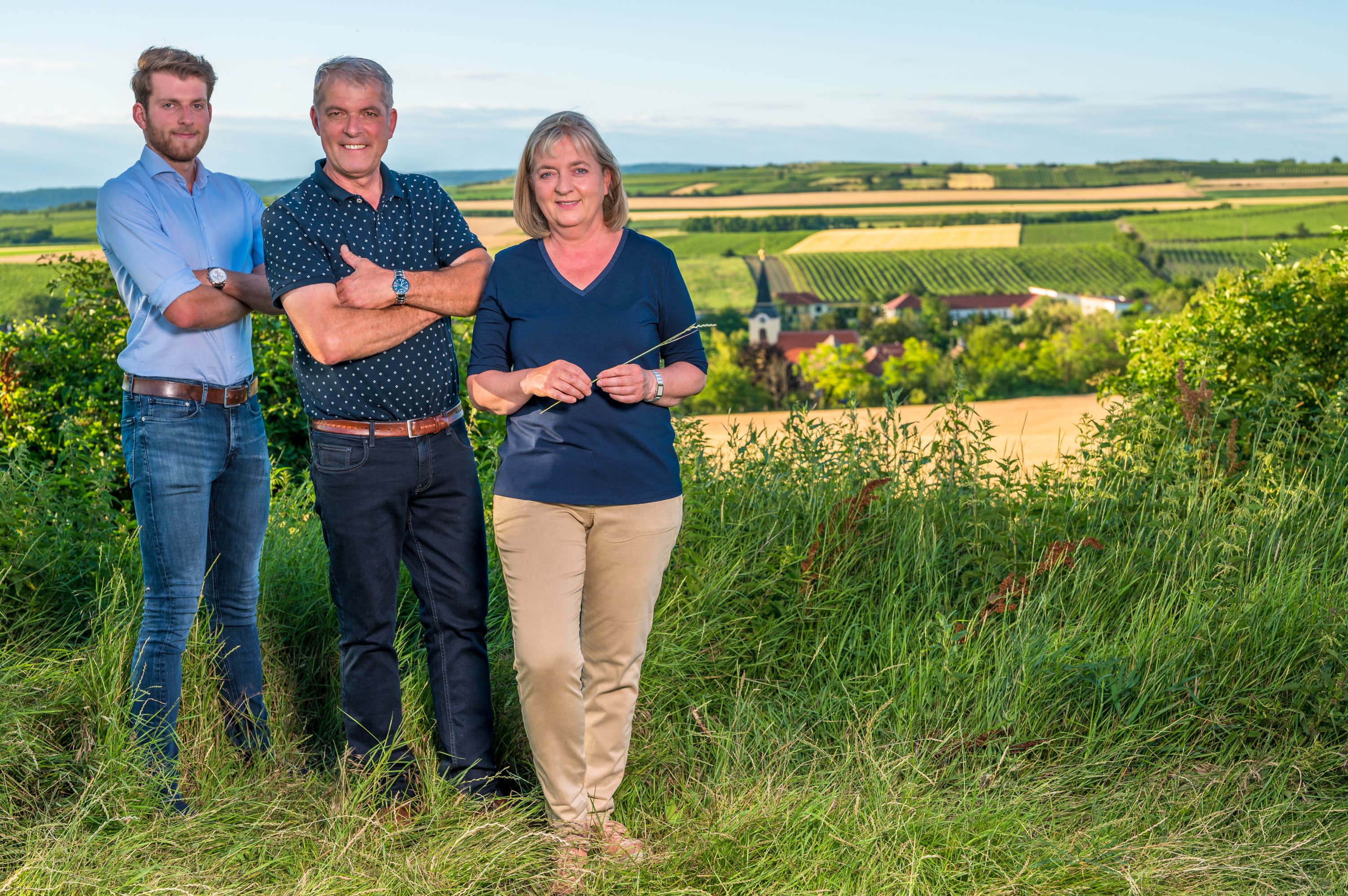 Három ember áll egy szőlőben, a háttérben egy vidéki tájra nyíló kilátással.