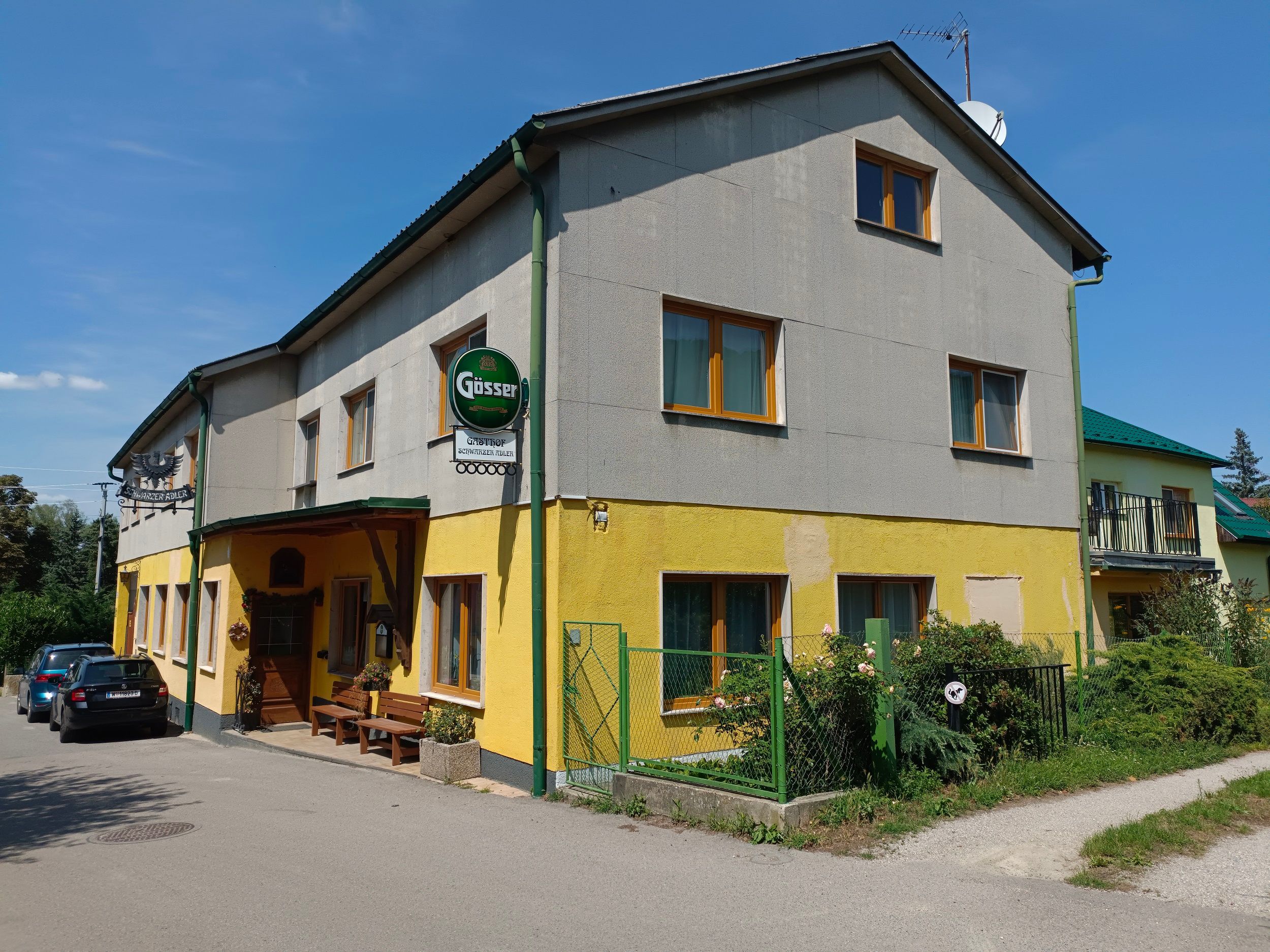 Egy sárga-szürke vendéglő külső nézete Gösser felirattal és parkoló autókkal.