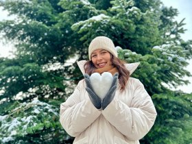Feh&eacute;r kab&aacute;tos nő egy h&oacute;b&oacute;l k&eacute;sz&uuml;lt sz&iacute;vet tart egy h&oacute;val bor&iacute;tott fa előtt.