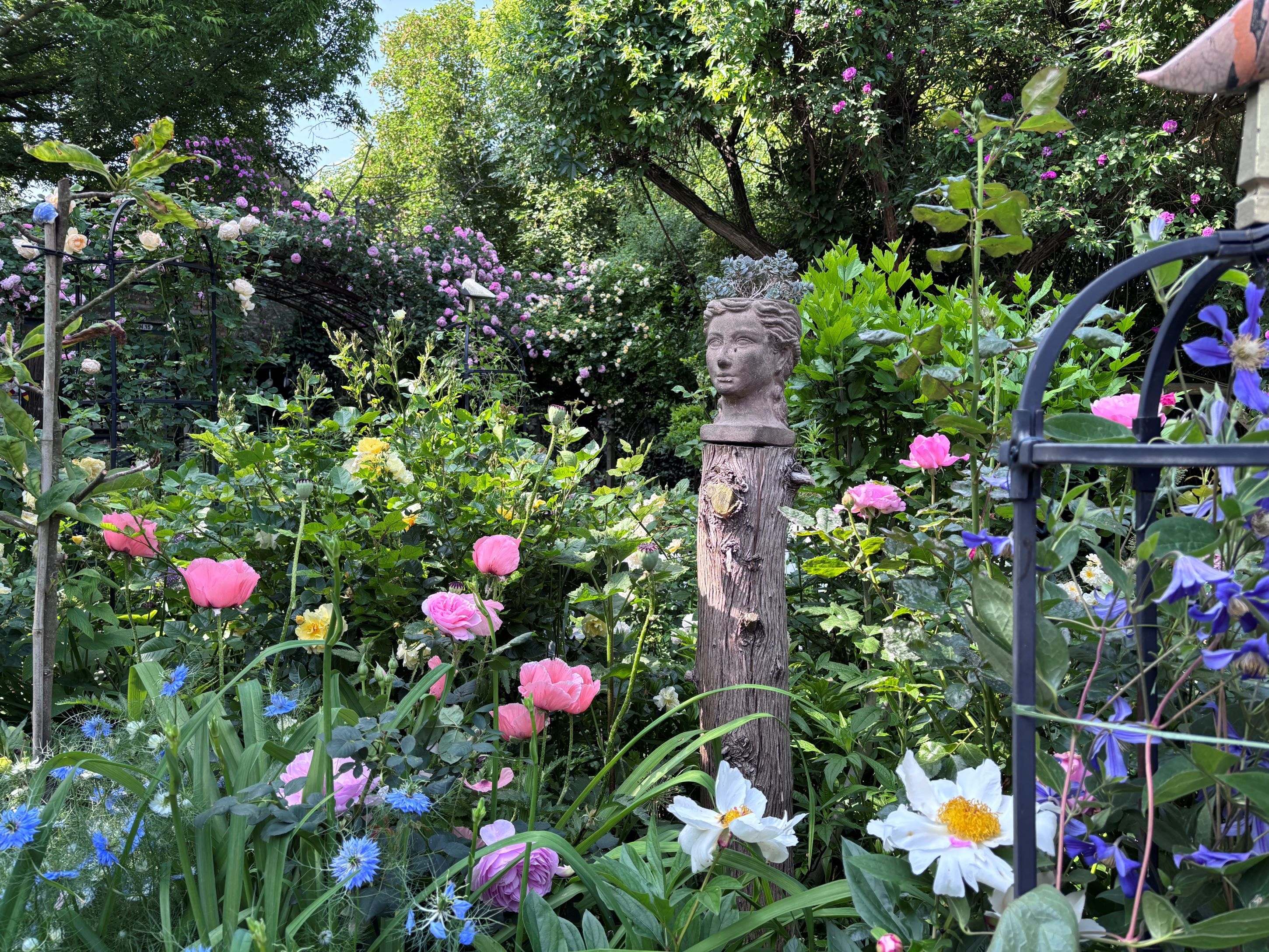 Virágzó kert színes virágokkal és egy mellszoborral egy faoszlopon.