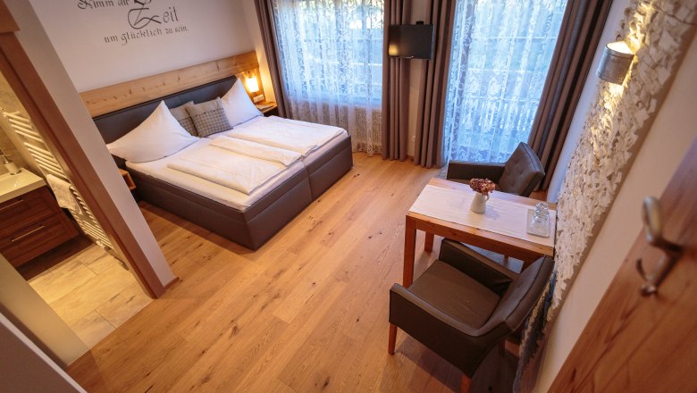 Hangulatos kétágyas szoba, fapadlóval, franciaággyal, kis asztallal és székekkel, dekoratív függönyökkel és TV-vel a falon.