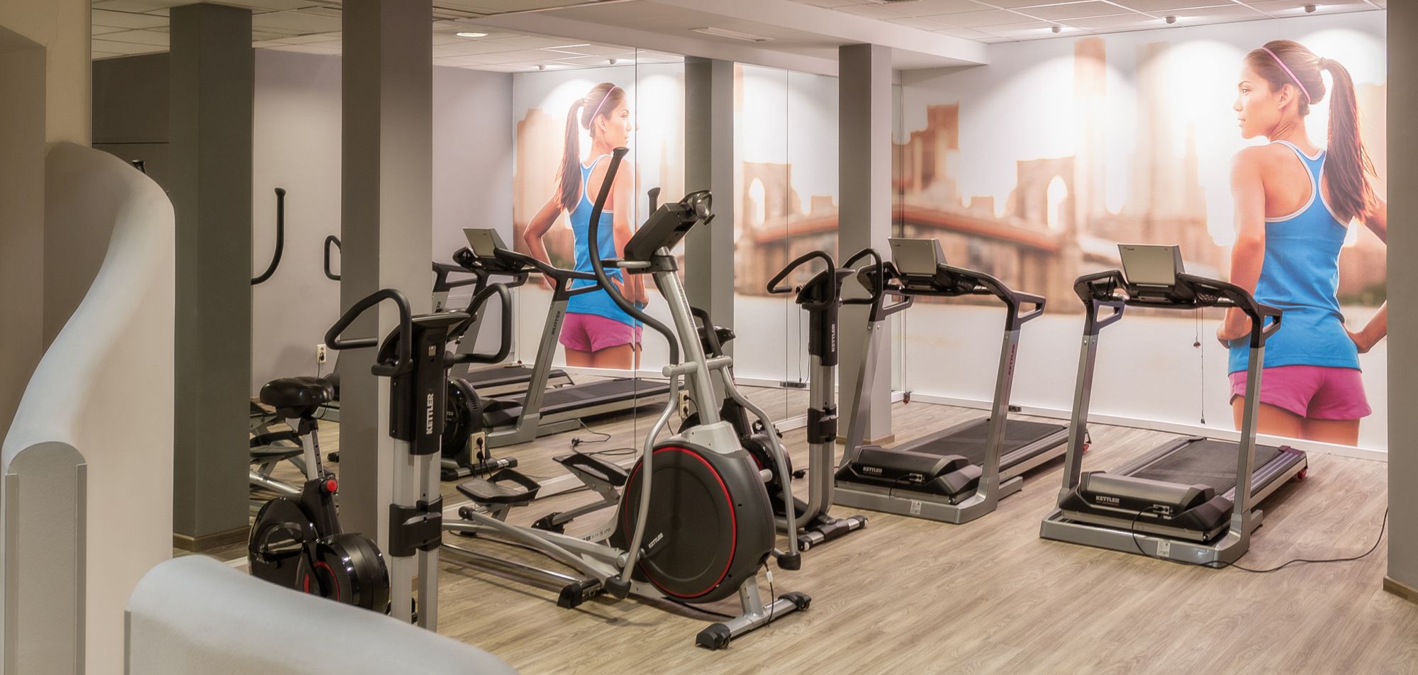 Fitneszterem futópadokkal és fitneszeszközökkel, falfestmény egy futó nőt ábrázol.