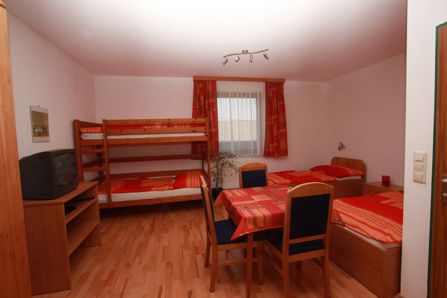 Egyszerűen berendezett szoba emeletes ággyal, egyszemélyes ággyal, asztallal és székekkel. Egy polcon televízió található. A háttérben függönyös ablak.