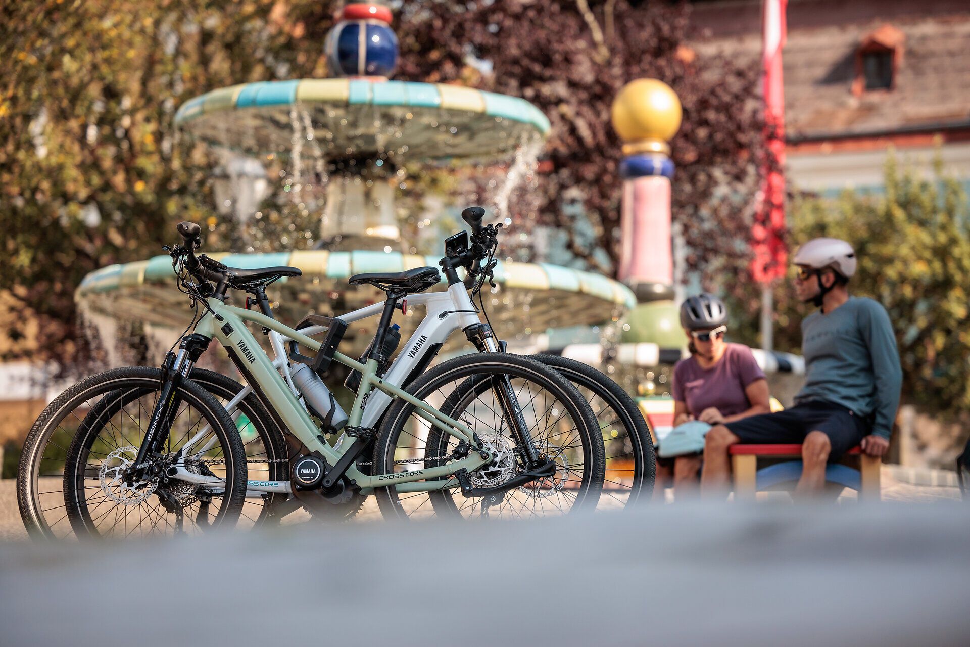 A kerékpárosok a meleg napsütésben pihennek a Hundertwasser-kútnál, amelyet pezsgő víz és színes növények vesznek körül. A friss levegő és a festői táj arra hívja Önt, hogy élvezze a természet szépségét és feltöltődjön.