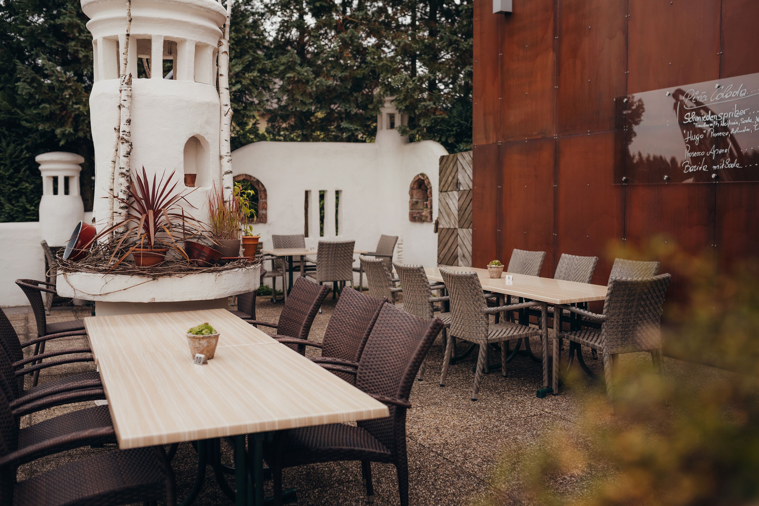 Hangulatos vendégkert faasztalokkal, fonott székekkel és dísznövényekkel egy fehér toronyban.