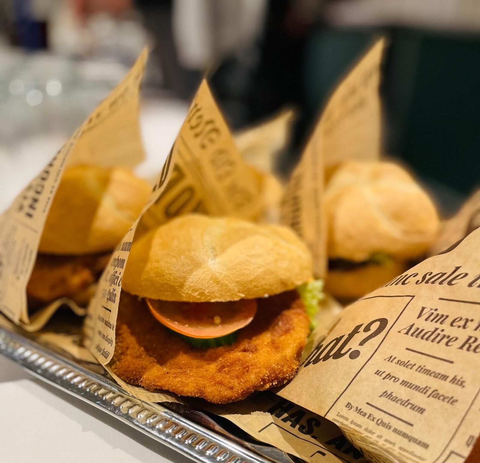 Több, újságpapírba csomagolt hamburger egy tálcán, az előtérben egy rántott húsos hamburger paradicsommal és uborkával.