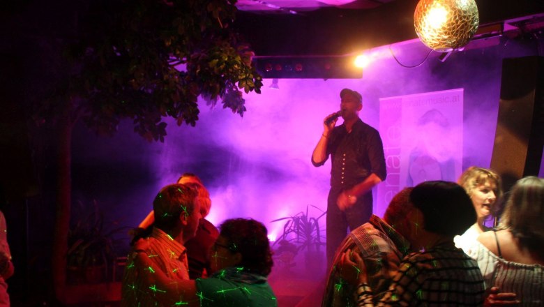 Egy &eacute;nekesnő a sz&iacute;npadon mikrofonnal, k&ouml;r&uuml;l&ouml;tte t&aacute;ncol&oacute; p&aacute;rok &eacute;s sz&iacute;nes f&eacute;nyek.