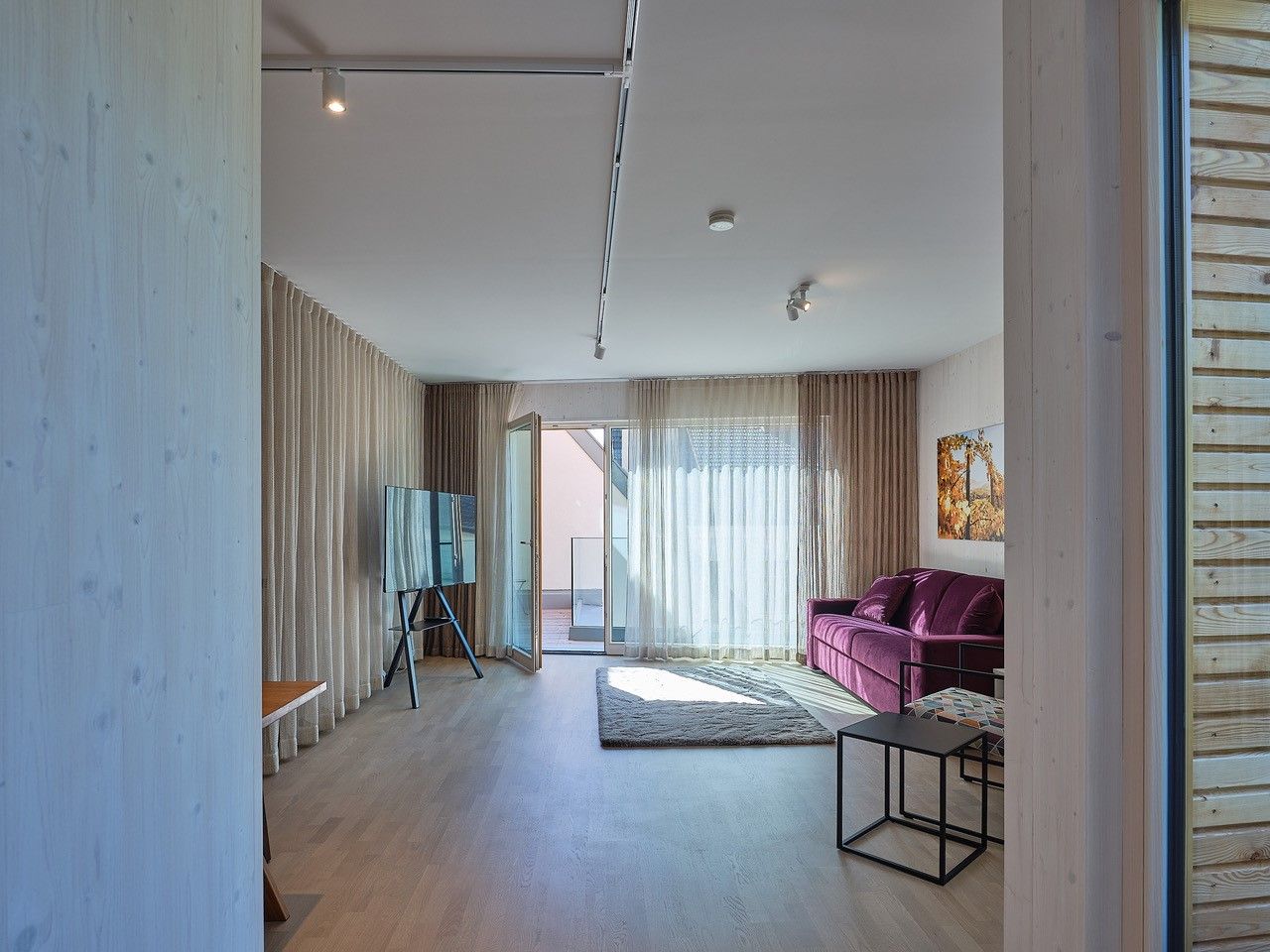 Modern nappali lila kanapéval, nagy ablakkal és állványon elhelyezett TV-vel.