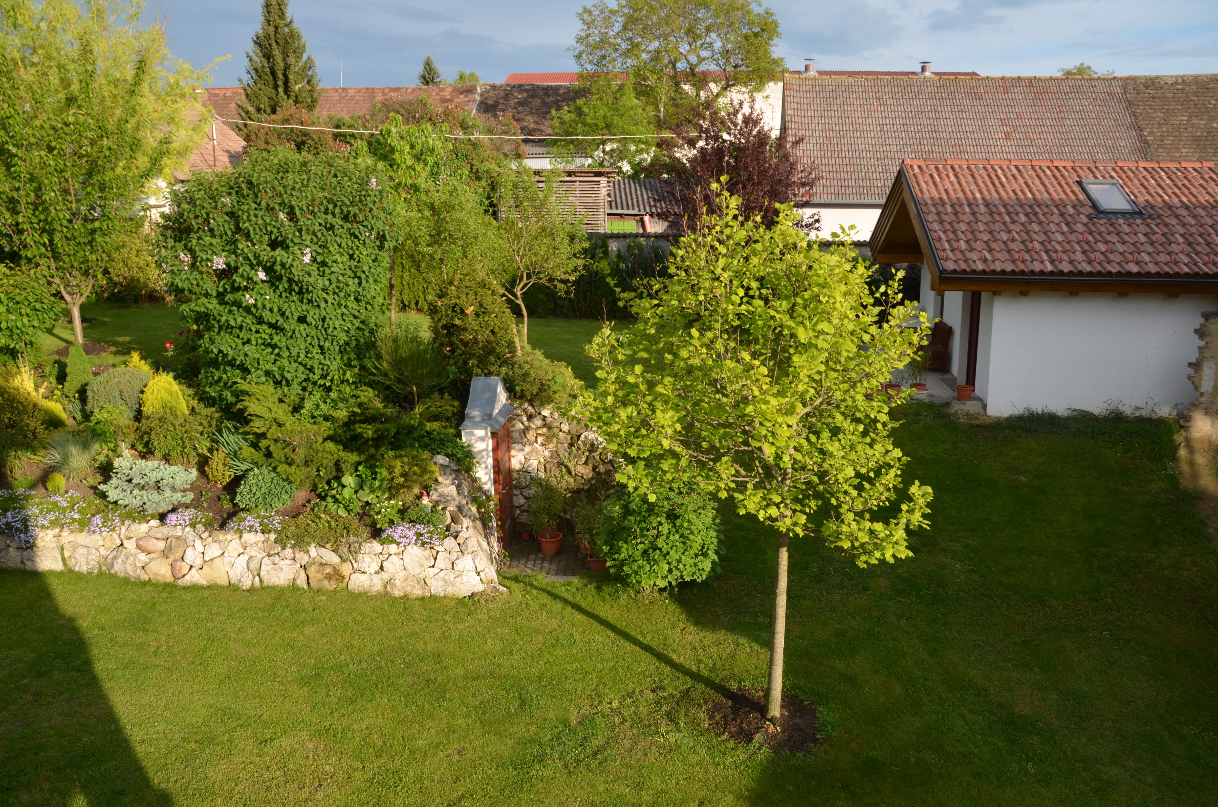 Gondozott kert fákkal, bokrokkal és egy kis cseréptetős épülettel.