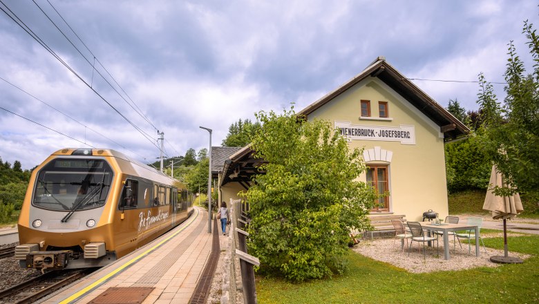 Ünnepek a BAHNhofban, © Niederösterreich Werbung / Maximilian Pawlikowsky A Wienerbruck-Josefsberg állomáson megáll egy aranyszínű vonat, amelyet zöld vidék és egy kis szabadtéri ülőhely vesz körül.