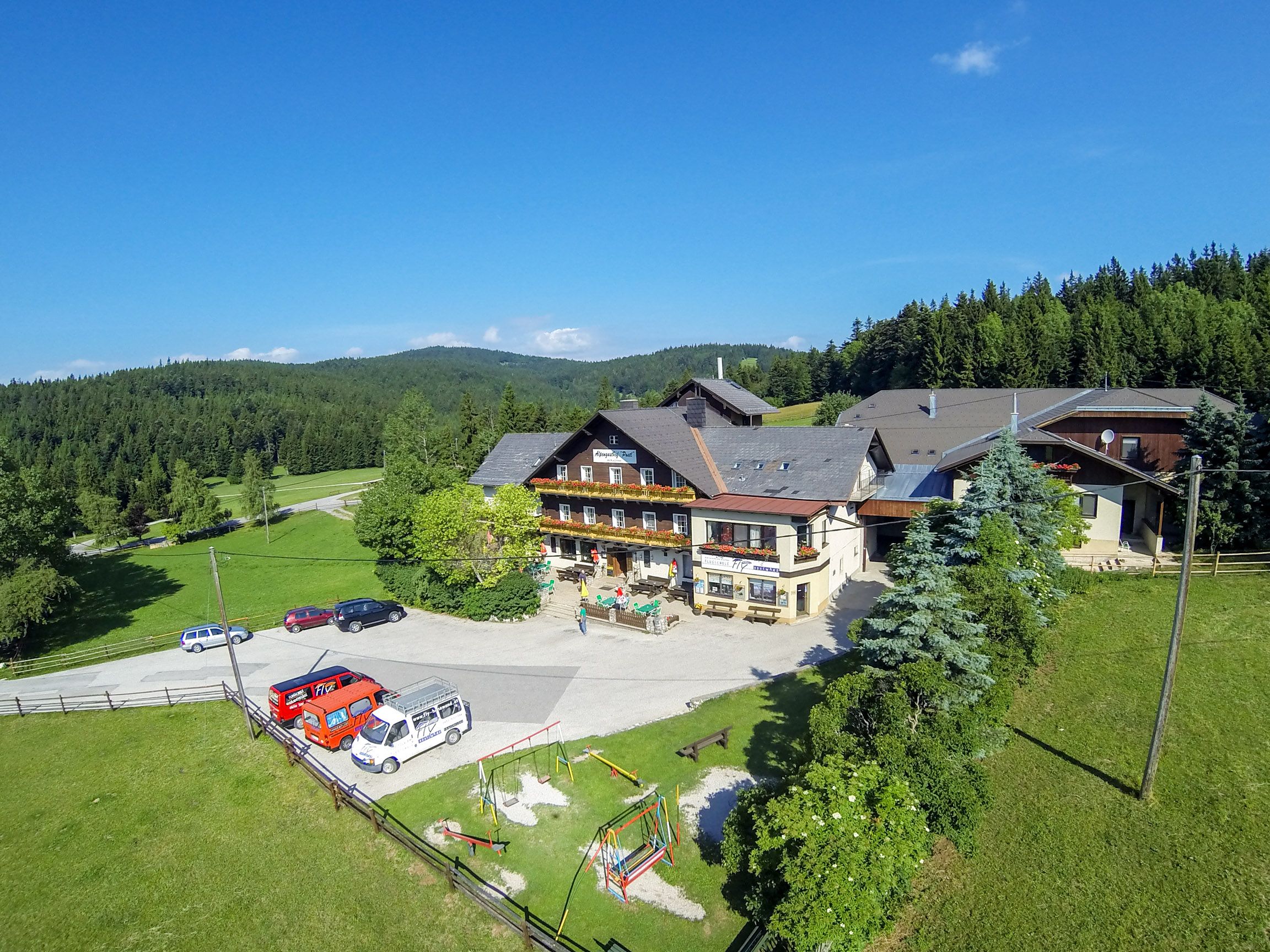 Alpengasthof Postl egy zöld, erdős tájban, előtérben több parkoló autóval és egy játszótérrel.