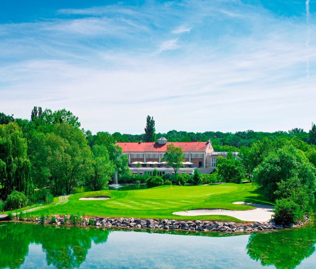 Golfpálya, a háttérben a klubházzal, fákkal és egy tóval körülvéve.