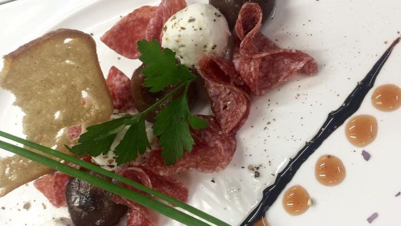 Egy tányér szalámival, kecskesajtgolyóval, fűszernövényekkel és egy szelet kenyérrel.