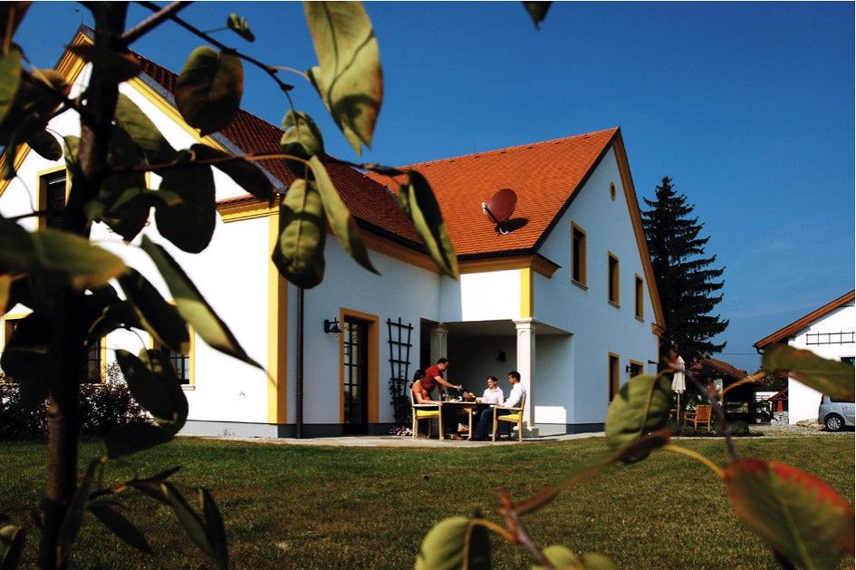 Fehér ház piros tetővel, zöld pázsittal és fákkal körülvéve. Odakint emberek ülnek egy asztalnál.