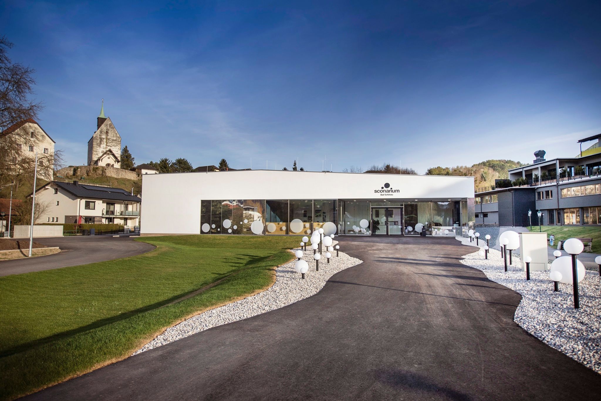 Modern Sconarium épület üvegfronttal és dekoratív gömblámpákkal, zöld területekkel körülvéve, a háttérben az erődtemplommal.