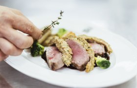 Közelkép egy rózsaszínű sült húsételről, fűszerkéreggel, brokkolival és egy kézzel kakukkfűvel.