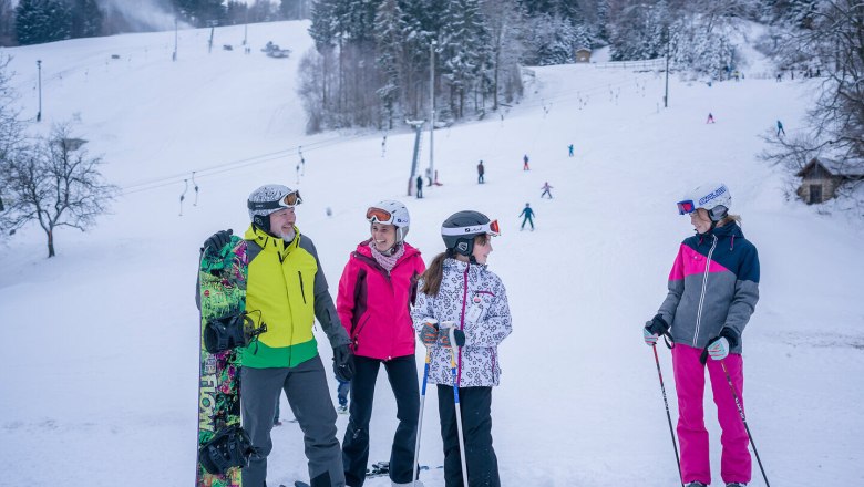 S&iacute;elők &eacute;s snowboardosok csoportja egy havas p&aacute;ly&aacute;n, a h&aacute;tt&eacute;rben erdő.