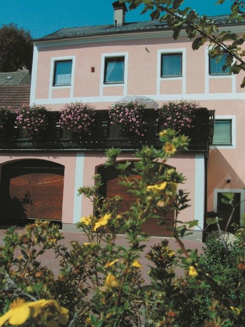 Egy kétszintes rózsaszín épület, virágzó növényekkel az erkélyen és sárga virágokkal az előtérben.