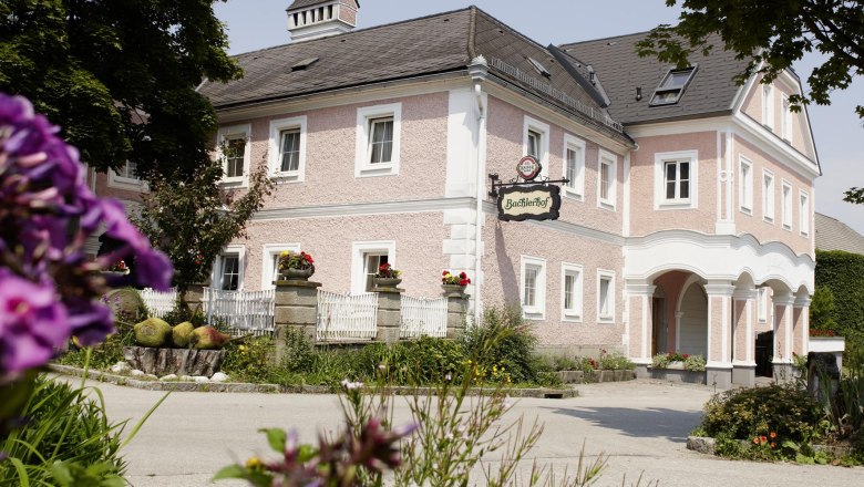 Hagyományos, rózsaszínűre festett épület a "Bachlerhof" felirattal, virágokkal és fákkal körülvéve.